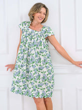 Load image into Gallery viewer, A woman wearing a short-sleeve white dress with a green and blue floral print, posing against a white paneled wall.
